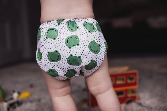 A small child wearing a frog print reusable cloth nappy walking away from the camera.