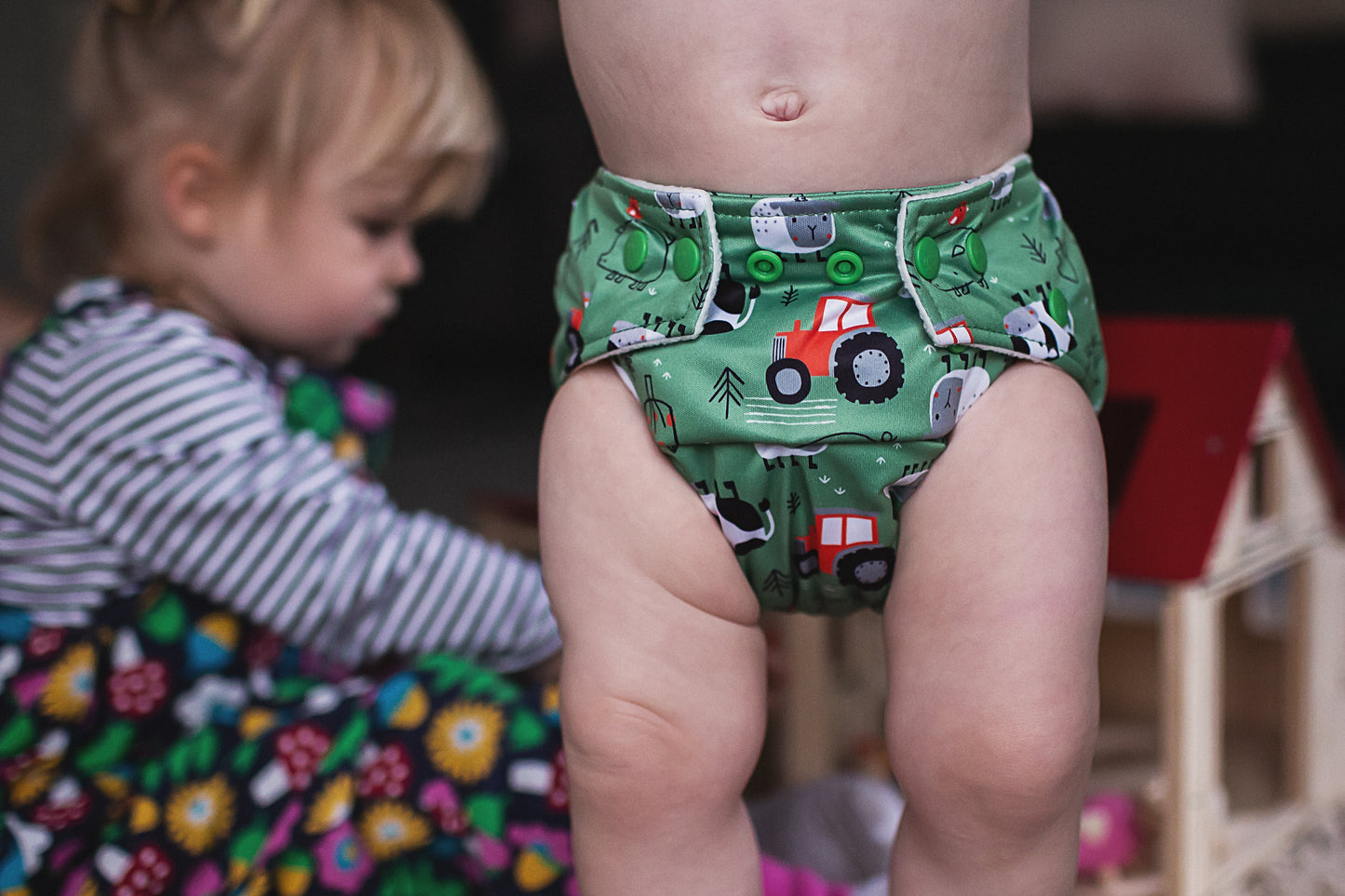 A small child wearing a green, farm print reusable cloth nappy. The child is standing up and there is another child playing with a wooden farm in the background. 