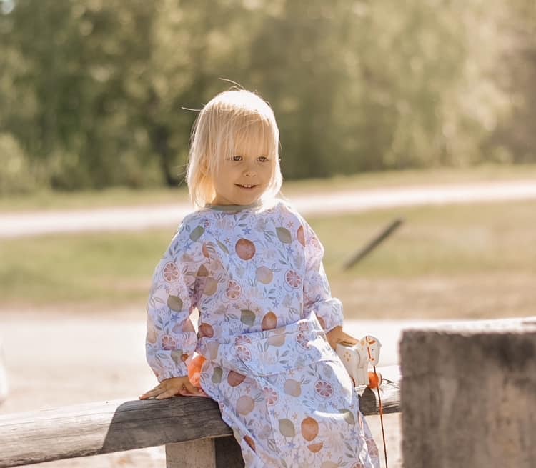 Image showing small child sitting on a fence wearing a shield coverall in 'when life gives you lemons' print.