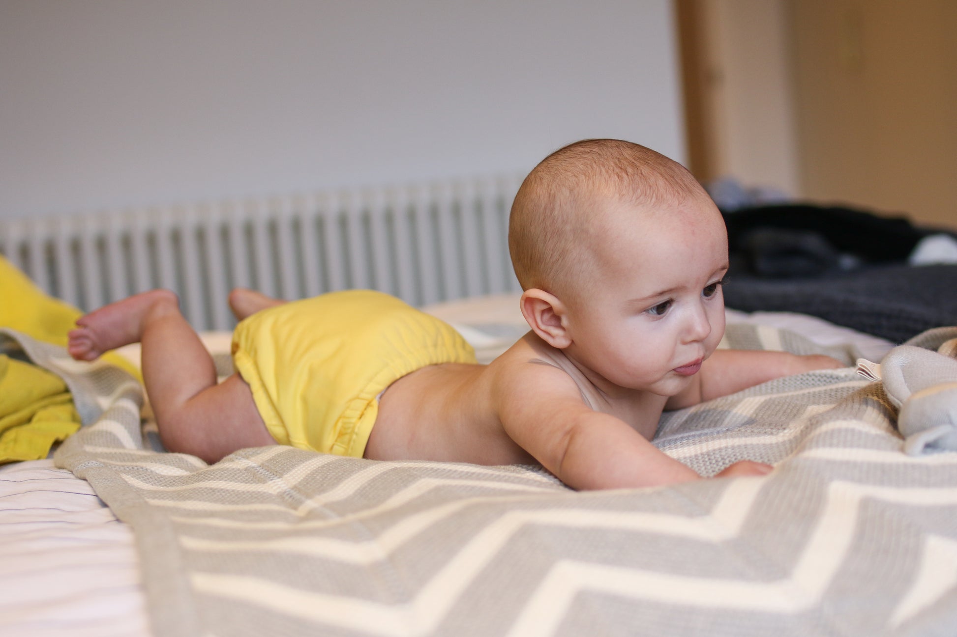 Image shows a young baby lying on their front on a bed wearing a yellow reusable cloth nappy. 