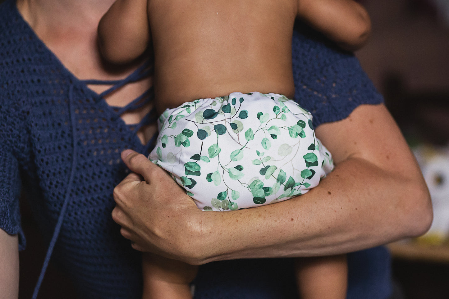 A small child being held by an adult. The child is wearing a reusable cloth nappy with a green leaf pattern. 