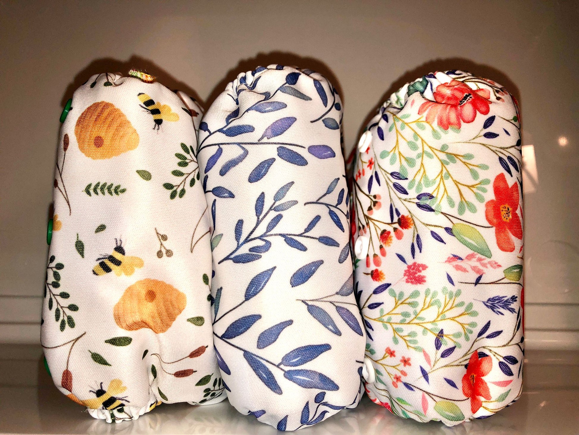 Image of three cloth nappies that come in this set. One with a bee print, one with a blue leaf print and one with a pink and blue flower print. 